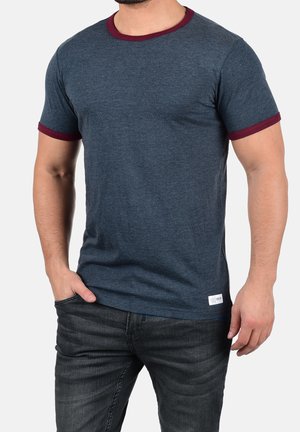 Homme portant un t-shirt bleu foncé à manches courtes avec un col et des bords de manches bordeaux, assorti à un jean noir, main dans la poche.