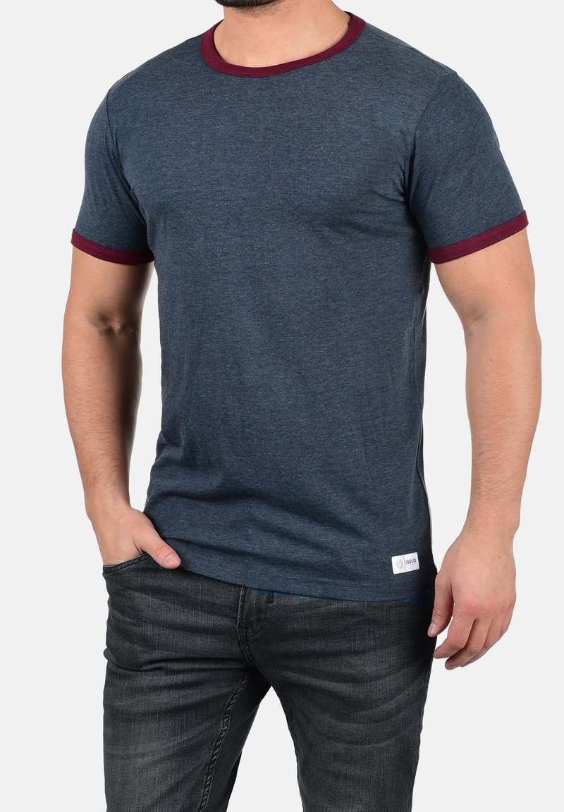 Homme portant un t-shirt bleu foncé à manches courtes avec un col et des bords de manches bordeaux, assorti à un jean noir, main dans la poche.