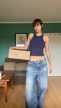 Jūrinės juostuotos marškinėlės be rankovių derinamos su laisvais šviesiai mėlynais džinsais. Modelis laiko rudą Zalando dėžutę. Paprasta fono spalva su žaliais sienų tonais.