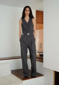 Grauer maßgeschneiderter Weste mit drei Knöpfen, kombiniert mit hoch taillierten, gefältelten grauen Hosen. Glatter Stoff und minimalistische Designdetails.