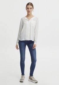 Blusa bianca a maniche lunghe con una chiusura a bottoni sul davanti e una singola tasca sul petto, abbinata a jeans skinny blu e scarpe da ginnastica grigie.