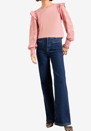 Femme portant un chemisier rose à manches longues bouffantes transparentes et un jean flare bleu foncé, associé à des chaussures pointues à imprimé léopard.