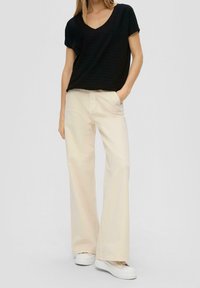 Žena v černém krátkém rukávu s výstřihem do V, béžových širokých kalhotách a béžových platformových teniskách, stojící s jednou rukou v kapse.