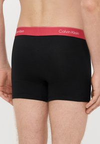 Musta bokserishortsit, joissa on punainen vyötärönauha ja "Calvin Klein" -logo. Kangas on sileä ja istuva, vakioleikkauksella eikä siinä ole näkyviä kuvioita.