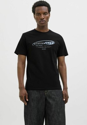 Schwarzes Baumwoll-T-Shirt mit grafischem Logo in Blau und Weiß, kurzen Ärmeln und klassischem Rundhalsausschnitt. Kombiniert mit dunklen Jeans.