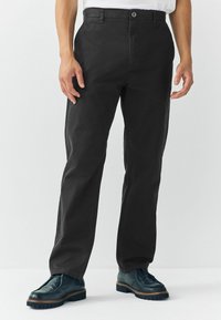 Man in zwarte rechtlopende broek en donkergroene veterschoenen, staand tegen een effen lichte achtergrond.