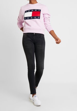 Henkilö pukeutuneena vaaleanpunaiseen Tommy Jeans -collegepaitaan, mustiin trikoihin ja valkoisiin Reebok-tossuihin kävelemässä valkoista taustaa vasten.