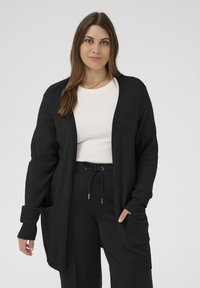 Cardigan noir ouvert sur le devant avec des poignets côtelés et des poches, fabriqué en tissu doux. Porté sur un haut blanc, associé à un pantalon noir.