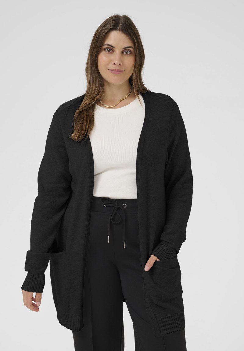 Cardigan noir ouvert sur le devant avec des poignets côtelés et des poches, fabriqué en tissu doux. Porté sur un haut blanc, associé à un pantalon noir.