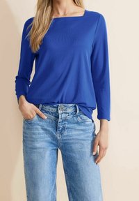 Camicia blu a maniche lunghe con scollatura quadrata, abbinata a jeans in denim blu chiaro. La camicia ha una texture liscia e una vestibilità rilassata.