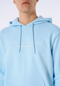 Felpa con cappuccio di colore azzurro chiaro, realizzata in tessuto morbido; presenta una tasca frontale e un testo in rilievo "COPENHAGEN LINDBERGH." Coulisse con punte blu.