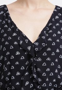 Svart blus med djup V-ringning, med vita triangulära mönster och en volangdetalj vid halsringningen. Mjuk, lättvikts tyg.