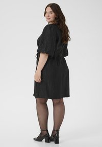 Vrouw met lang bruin haar, gekleed in een getextureerde zwarte jurk tot de knie, doorschijnende zwarte panty en zwarte block-heeled schoenen, iets gekeerdstaand.