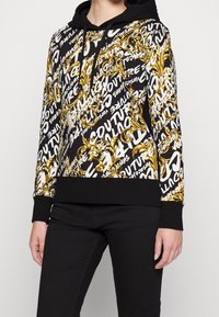 Mikina s kapucí, která má zlatý a černý ornamentální vzor s bílými textovými akcenty. Materiál je látkový, s vroubkovanými černými manžetami a dolním lemem.
