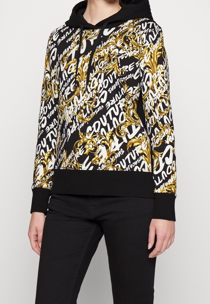Mikina s kapucí, která má zlatý a černý ornamentální vzor s bílými textovými akcenty. Materiál je látkový, s vroubkovanými černými manžetami a dolním lemem.