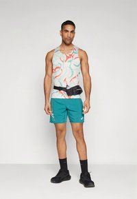 Vaaleansävyinen hihaton toppi, jossa abstrakteja punaisia ja sinisiä kuvioita; teepparin väriset shortsit sivulogoilla; musta vyölaukku; mustat lenkkarit.