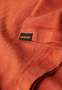 Oranje stof met een subtiele textuur en plooien. Zwarte label met "SUPERDRY" in oranje letters aan de zijkant. Zacht materiaal, casual ontwerp.