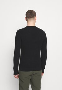 Schwarzer gerippter Pullover mit langen Ärmeln, einem runden Ausschnitt und einer taillierten Passform, kombiniert mit olivgrünen Hosen.