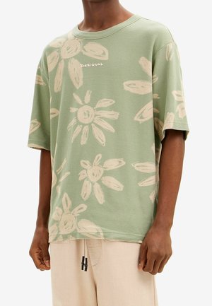 Homme portant un t-shirt vert ample avec des motifs floraux abstraits beige clair et le logo "Desigual", associé à un pantalon beige clair.