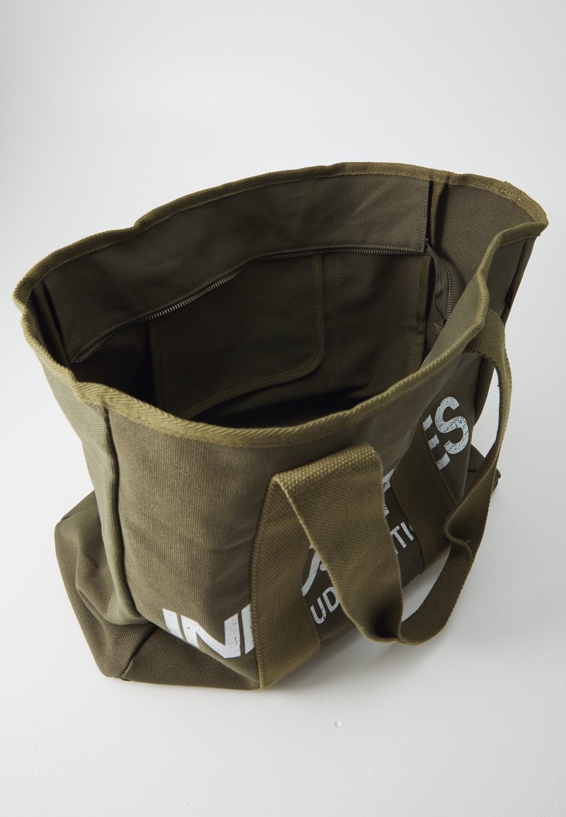 Oliwkowa torba na zakupy z płótna z dwoma mocnymi uchwytami, wewnętrzną kieszenią na zamek i białym nadrukowanym logo z przodu. Projekt z otwartym wierzchem.