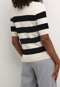 Personne portant un haut à rayures horizontales noir et blanc à manches courtes et un pantalon gris clair, se tenant devant un mur clair et uni.