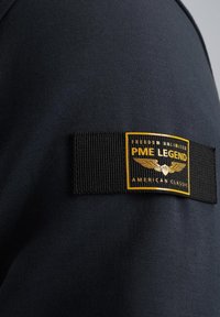 Sort t-shirt lavet af struktureret stof, med en rektangulær label der viser "PME LEGEND" i guld og sorte farver samt luftfartsinspirerede detaljer.