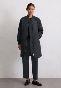 Femme aux cheveux foncés attachés, portant un long manteau noir matelassé, un pantalon sombre, des mocassins noirs, se tenant contre un mur clair.
