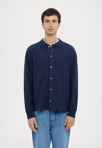 Camicia a maniche lunghe blu navy con bottoni, realizzata in tessuto morbido, dotata di colletto, bottoni frontali e polsini a costine. Indossata con jeans in denim blu chiaro.