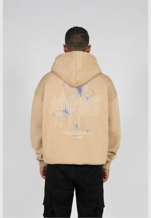 Homme vu de dos portant un sweat à capuche beige avec un motif de papillon violet clair et un texte blanc ondulé dans le dos, associé à un pantalon cargo noir.