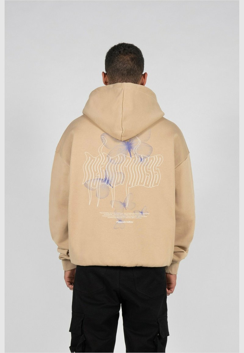 Homme vu de dos portant un sweat à capuche beige avec un motif de papillon violet clair et un texte blanc ondulé dans le dos, associé à un pantalon cargo noir.