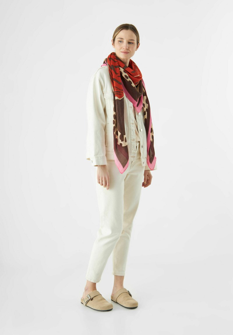 Grande écharpe à motifs dans des tons chauds de rouge, brun et rose. portée sur une veste en denim de couleur claire et un pantalon, créant un look superposé.