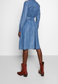 Denimklänning med lång ärm, åtdragen midja och flödande kjol. Har krage, knappmanschetter och råkant. Matchad med bruna anklarstövlar.