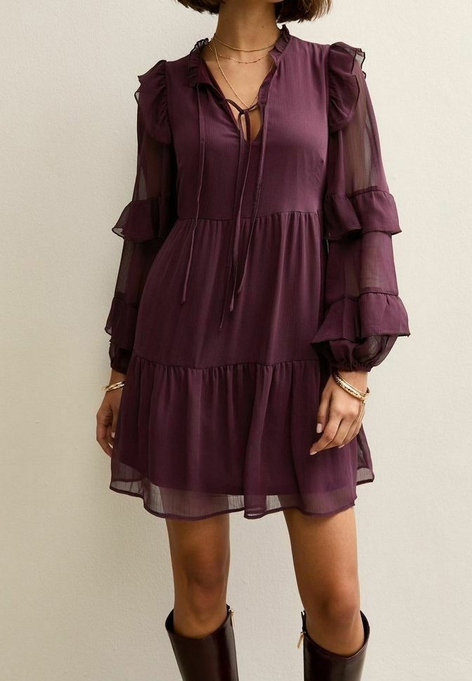 Femme portant une robe prune violette mi-longue avec des manches longues bouffantes transparentes et des détails à volants, associée à des bottes hautes marron foncé.