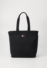 Svart tote-bag laget av lerret, med rektangulær form, doble stoffhåndtak og et lite logo-patch i rød, hvit og blå.