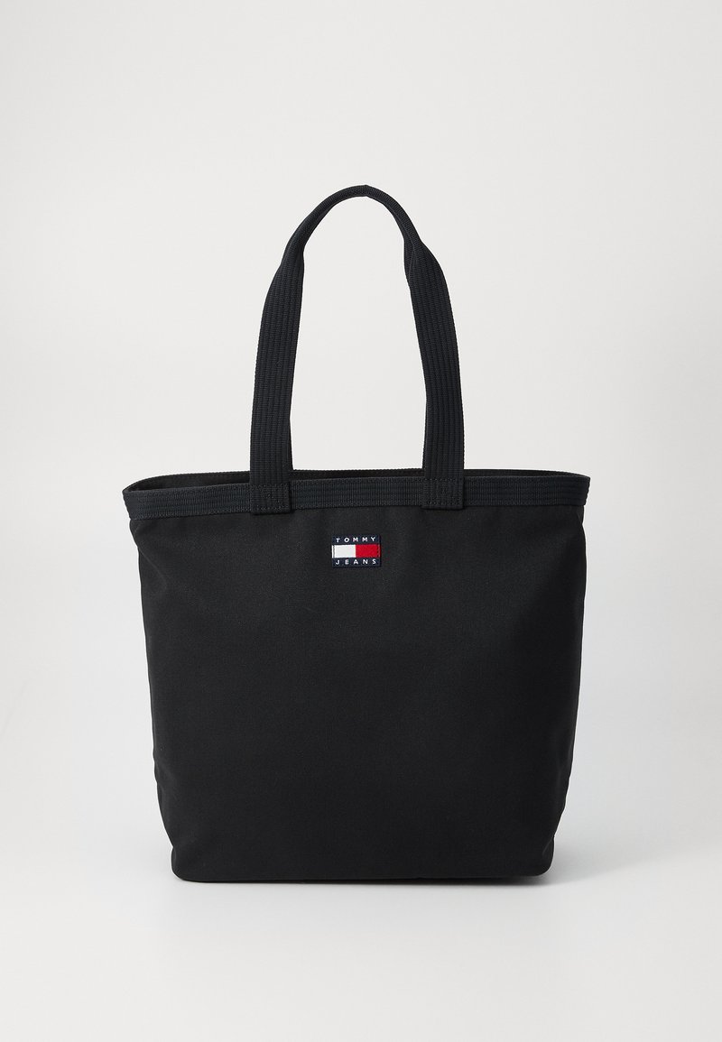 Juodas drobės pirkinių krepšys, turintis stačiakampę formą, dvi medžiaginias rankenas ir mažą logotipo pleistrą raudonai, baltai ir mėlynai.