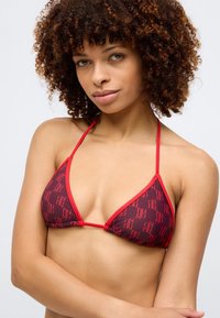 Raudona trikampė bikini viršūnė su plonais petnešomis ir logotipo raštu. Lygi medžiaga su kontrastuojančiomis raudonomis detalėmis ir minimalistiniu dizainu.