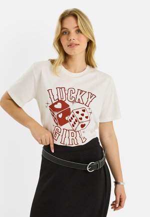 Mujer rubia con una camiseta blanca que dice "Lucky Girl" con dados rojos y corazones, combinada con una falda negra y un cinturón.