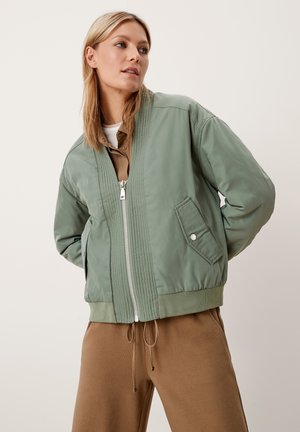 Jonge vrouw met licht haar draagt een saliegroene hoodie over brown trekkoordbroek en kijkt lichtjes opzij tegen een effen achtergrond.
