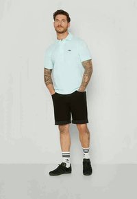 Mies, jolla on tatuointeja, pukeutuneena vaaleansiniseen polo-paitaan, mustiin lyhyisiin shortseihin, valkoisiin Adidas-sukkiin ja mustiin tennarit, seisoo kädet taskuissa.