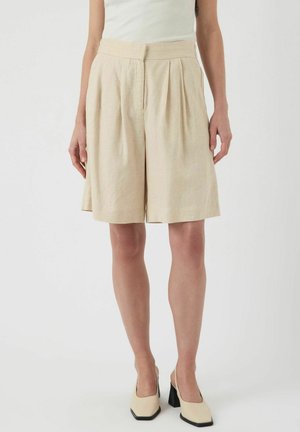 Pantalones cortos de lino beige con un frente plisado, piernas anchas y una cintura alta. Textura suave, bordes rematados y combinados con zapatos de tacón color crema.