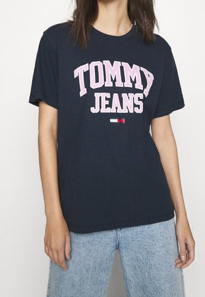 Person iført navyblå T-shirt med teksten "TOMMY JEANS" og logo, kombineret med lyseblå denimjeans mod en ensfarvet baggrund.