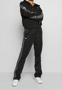 Survêtement de sport noir avec un sweat à capuche zippé, doté de bandes latérales blanches et d'accents de logo, accompagné d'un pantalon assorti. Finition en tissu texturé.