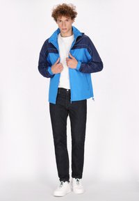 Blauwe en marinekleurige color-blocked jas met een rits aan de voorkant, verstelbare manchetten en een staande kraag, gecombineerd met donkere spijkerbroek en witte sneakers.