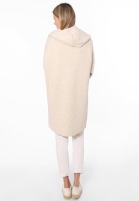 Beige, strukturierter, übergroßer Hoodie mit abgerundetem Saum und seitlichen Schlitzen, kombiniert mit cremefarbenen, körperbetonten Hosen und hellen Slippern.