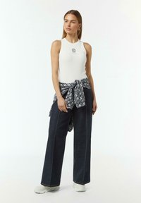 Witte geribde tanktop met een ronde halslijn, gecombineerd met een wijde donkere spijkerbroek en een blauw patroongevoel sjaal om de taille geknoopt. Witte sneakers.