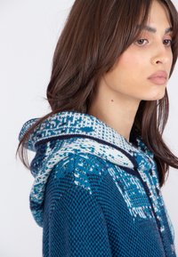 Gebreide hoodie in levendige blauwe tinten, met een gebloemde capuchon in wit en marineblauw. Zachte textuur met geometrische ontwerpelementen.