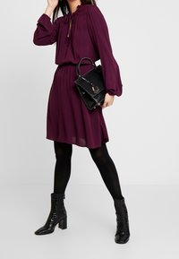 Lila klänning med dragsko runt nacken, långa ärmar och en utsvängd kjol. Matchad med en svart, texturerad handväska och glänsande svarta ankelstövlar.