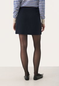 Mini-jupe marine avec une texture lisse, associée à des collants noirs transparents et des chaussures noires plates, vue de dos. Haut rayé partiellement visible.