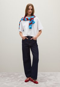 Witte oversized t-shirt, donkerblauwe wijde broek, rode ballerina's; blauw en rood gestreepte sjaal om de schouders gedrapeerd. Neutrale achtergrond.