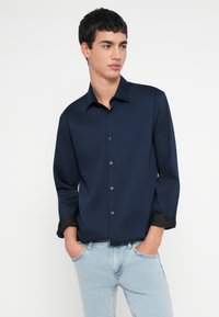 Tumesinine nööpidega särk kraega ja pikkade varrukatega, millel on mustad mansetid. Sile kangas ja minimalistlik disain.